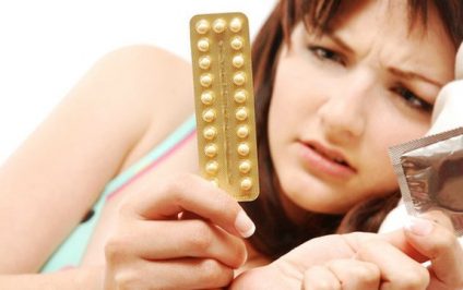 A beautiful young woman lying in bed confused about contraception. She is looking at contraptive pills and condoms and can't decide.